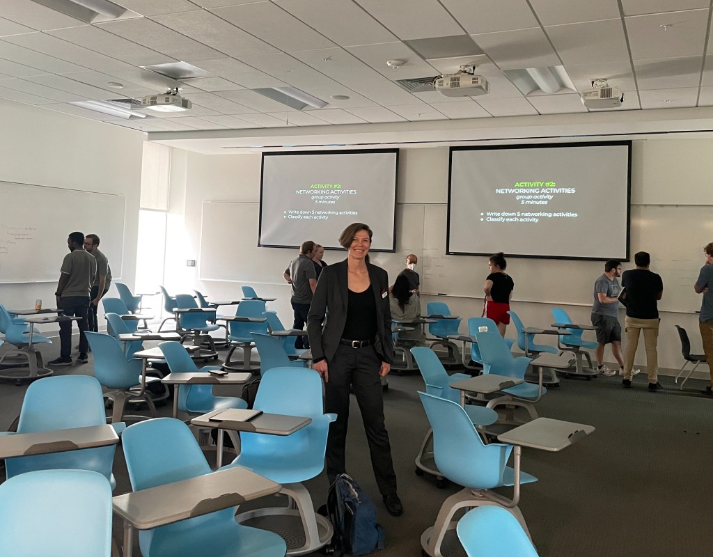 Christina stands surrounded by gray desks and blue and gray chairs. She is wearing an all-black outfit, and presenting screens and attendees are behind her.