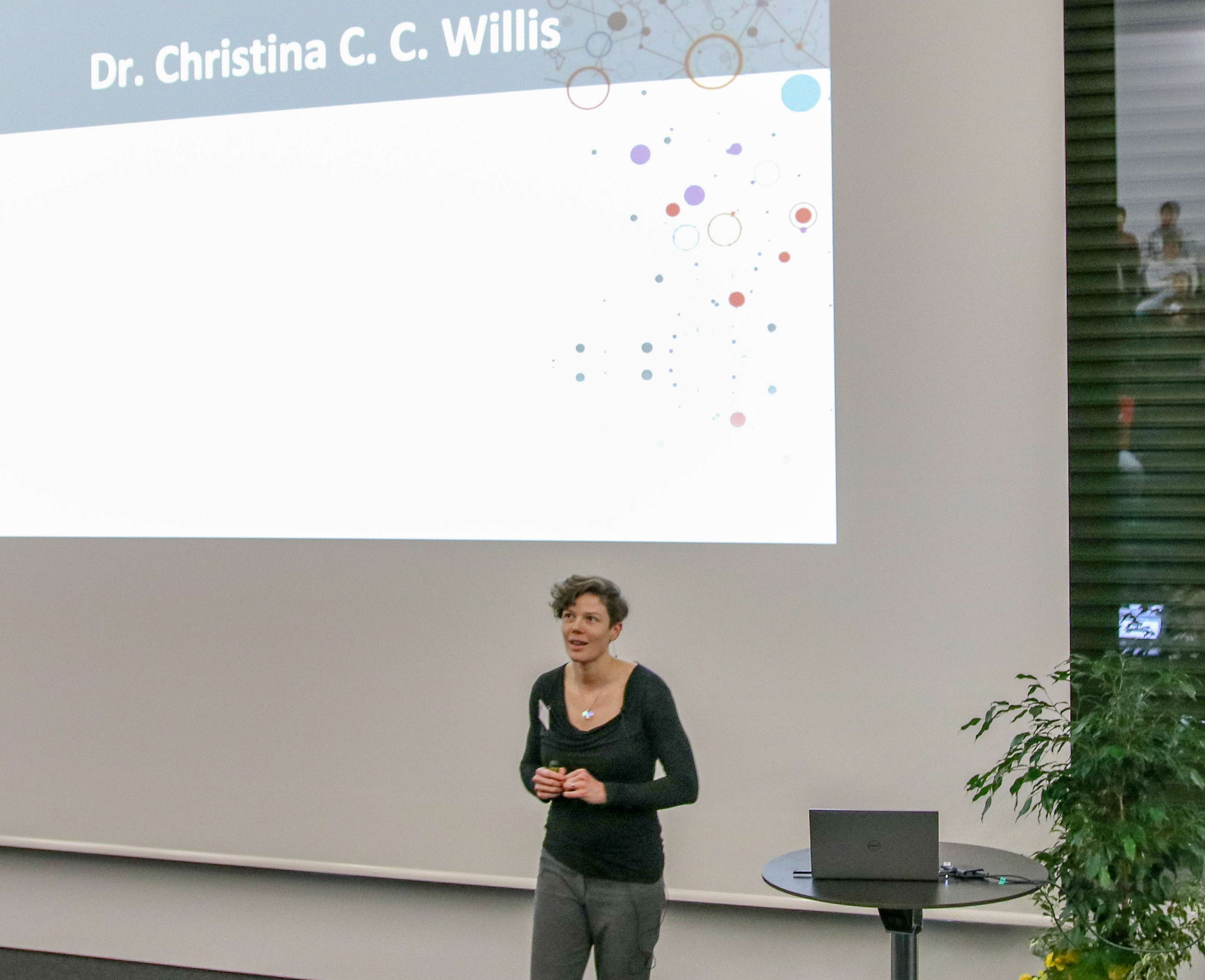 Christina stands in an auditorium weaking a black shirt and giving a lecture.