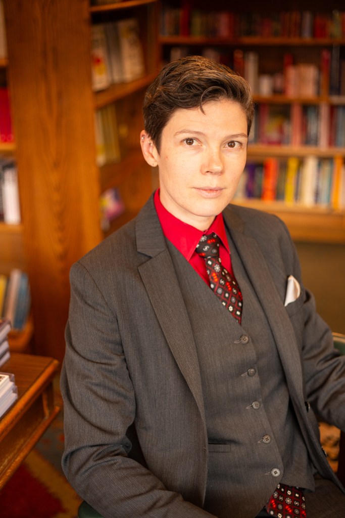 Christina sits in front of a bookshelf wearing a grey suit, red shirt, and black tie.
