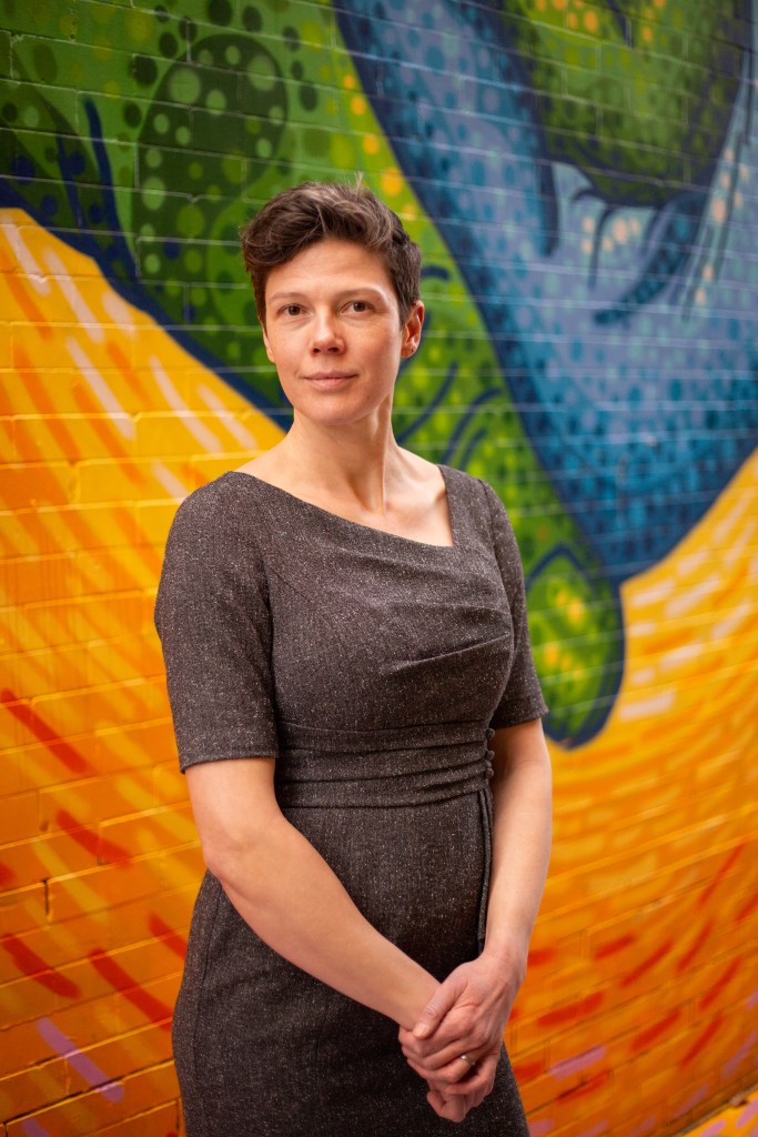 Christina stands against a colorful, abstract mural wearing a brown dress.